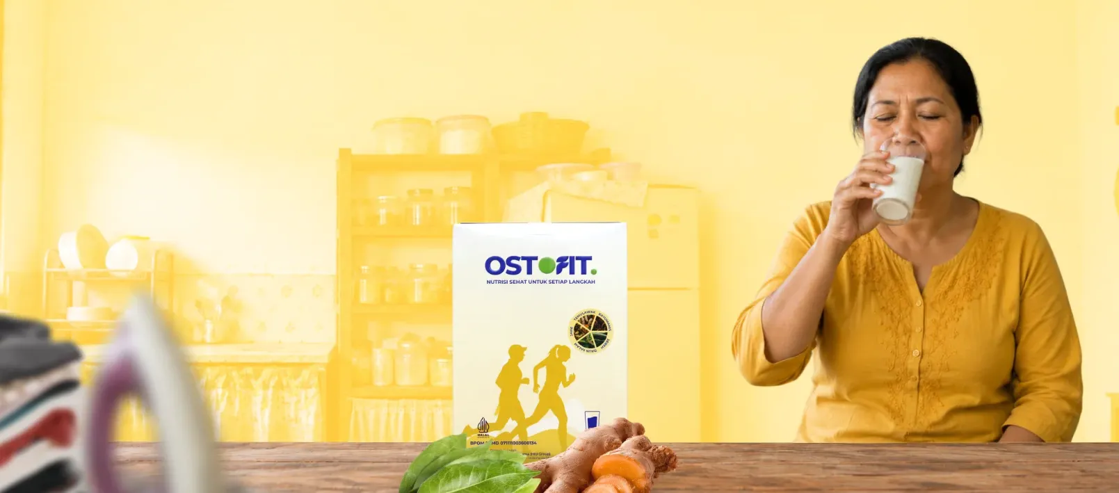 Seorang ibu rumah tangga meminum susu herbal di dapur, dengan kemasan produk Ostofit bertuliskan 'Nutrisi Sehat Untuk Setiap Langkah' di depannya bersama rempah alami.