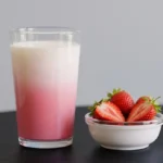 Segelas susu rasa stroberi dengan semangkuk buah stroberi yang telah dibelah.